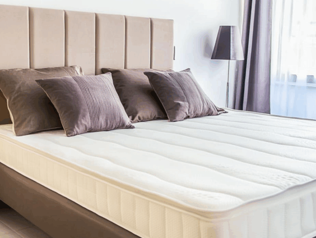 Ein ordentlich gemachtes Bett mit einem hellbeigen gepolsterten Kopfteil, vier braune Kissen, eine weiße Matratze zum Wechseln und eine Stehlampe neben einem Fenster mit durchsichtigen Vorhängen. Das Zimmer hat ein modernes, minimalistisches Design.