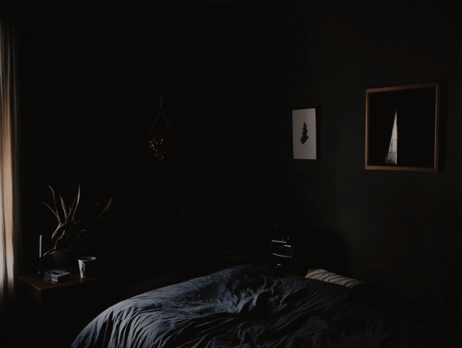 Ein schwach beleuchtetes Schlafzimmer mit dunklen Wänden, einem ungemachten Bett, einem Nachttisch mit einer Pflanze und einer Tasse sowie zwei gerahmten Bildern, die über dem Bett hängen, schafft eine Atmosphäre, die an einen Albtraum erinnert. Durch ein Fenster auf der linken Seite fällt sanftes Licht ein. Eine Situation, wo jemand gerade aus einem Albtraum erwacht sein könnte