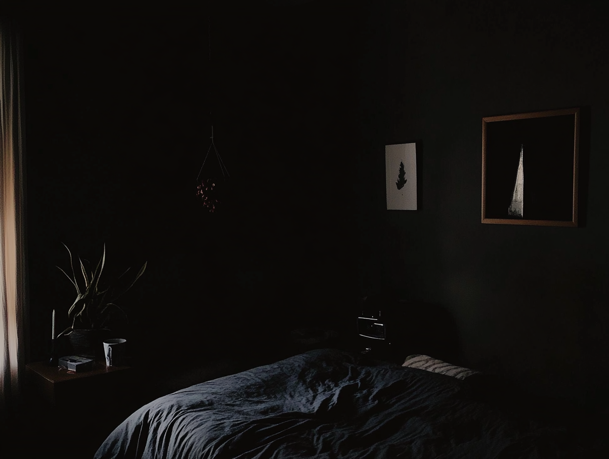 Ein schwach beleuchtetes Schlafzimmer mit dunklen Wänden, einem ungemachten Bett, einem Nachttisch mit einer Pflanze und einer Tasse sowie zwei gerahmten Bildern, die über dem Bett hängen, schafft eine Atmosphäre, die an einen Albtraum erinnert. Durch ein Fenster auf der linken Seite fällt sanftes Licht ein. Eine Situation, wo jemand gerade aus einem Albtraum erwacht sein könnte