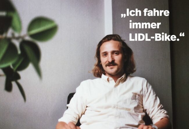 Ein Mann mit schulterlangem Haar und Schnurrbart, der einen hellen Button-up trägt, sitzt lächelnd auf einem Stuhl. Der deutsche Text lautet: "Ich fahre immer LIDL-Bike." Eine Blattpflanze steht unscharf im Vordergrund - perfekt für den kreativen Raum eines Grafikdesigners.