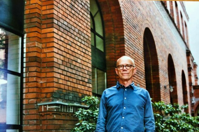 Ein Mann mit Brille und blauem Hemd steht vor einem Backsteingebäude mit großen Rundbogenfenstern, umgeben von Grünpflanzen - das erinnert an ein Interview mit Erik Spiekermann.