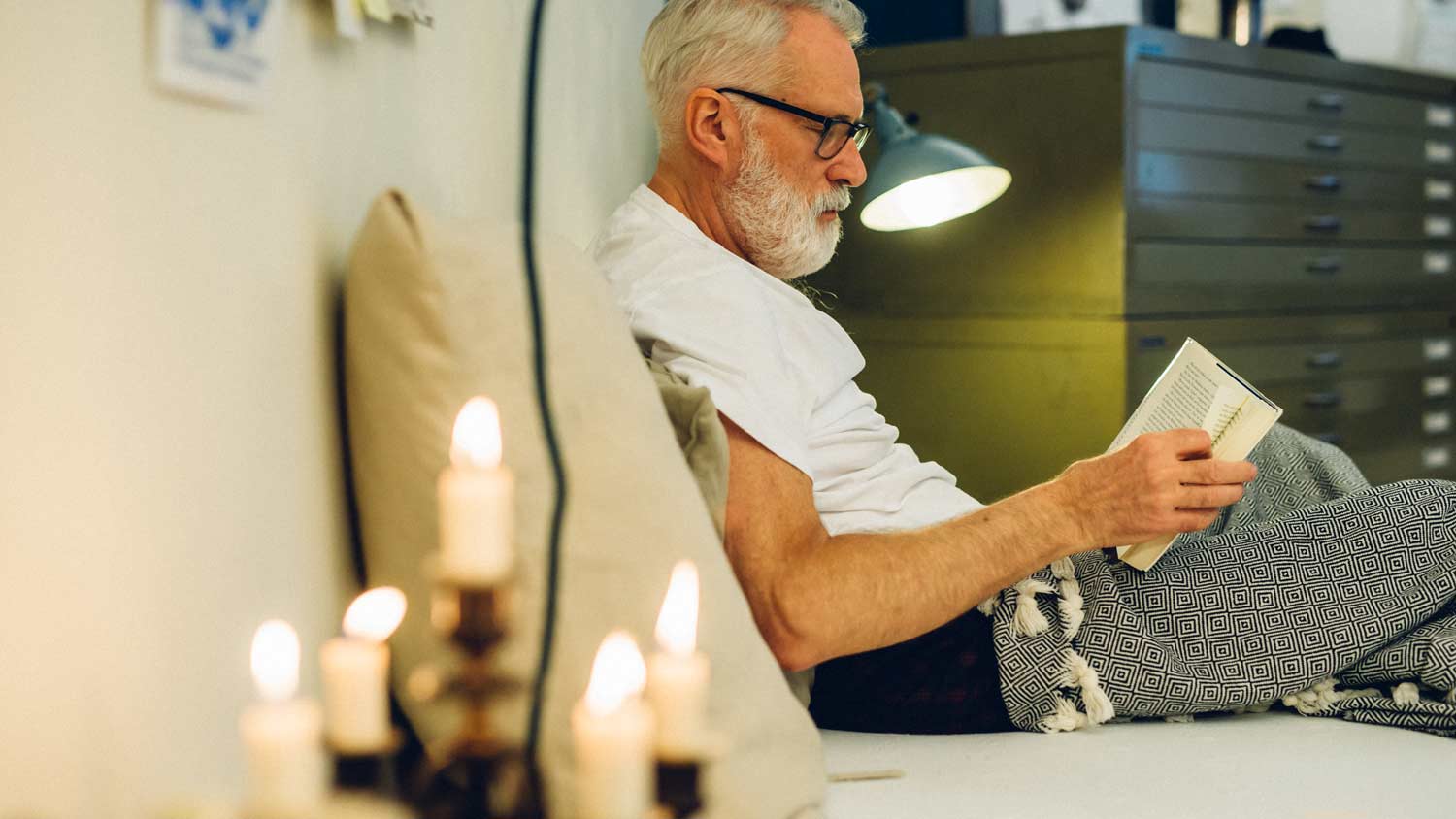 Ein älterer Mann mit Brille und weißem Bart liest ein Buch, während er auf einem Bett sitzt, das mit einer gemusterten Decke bedeckt ist. Auf einem Tisch im Vordergrund stehen brennende Kerzen, die eine gemütliche Atmosphäre schaffen.