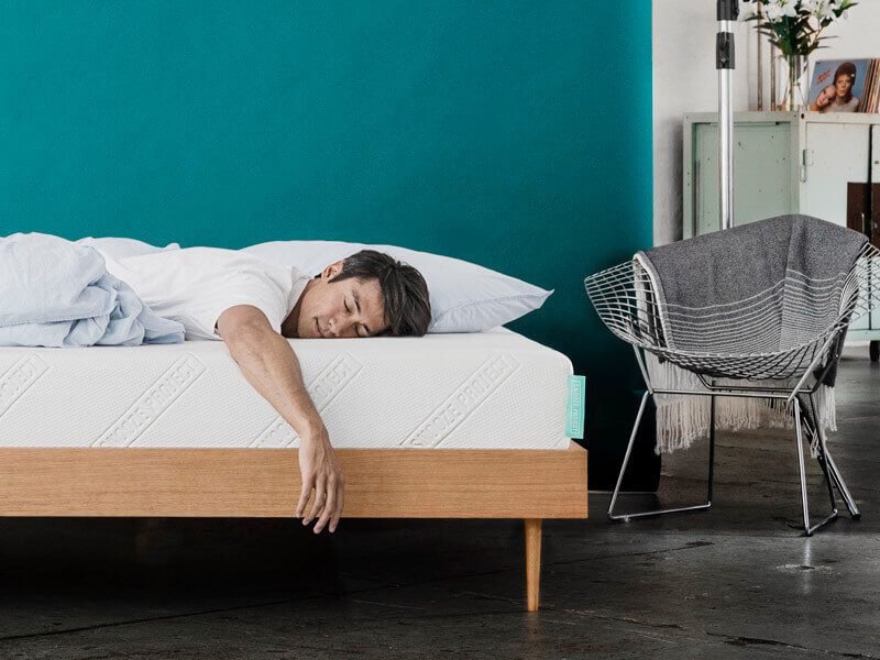 Ein Mann liegt mit dem Gesicht nach unten auf einem Bett mit weißer Matratze und hölzernem Rahmen, ein Arm hängt an der Seite herunter. Das Zimmer hat tealfarbene Wände, einen Stuhl mit einer grauen Decke und einige Regale und Dekorationen im Hintergrund.