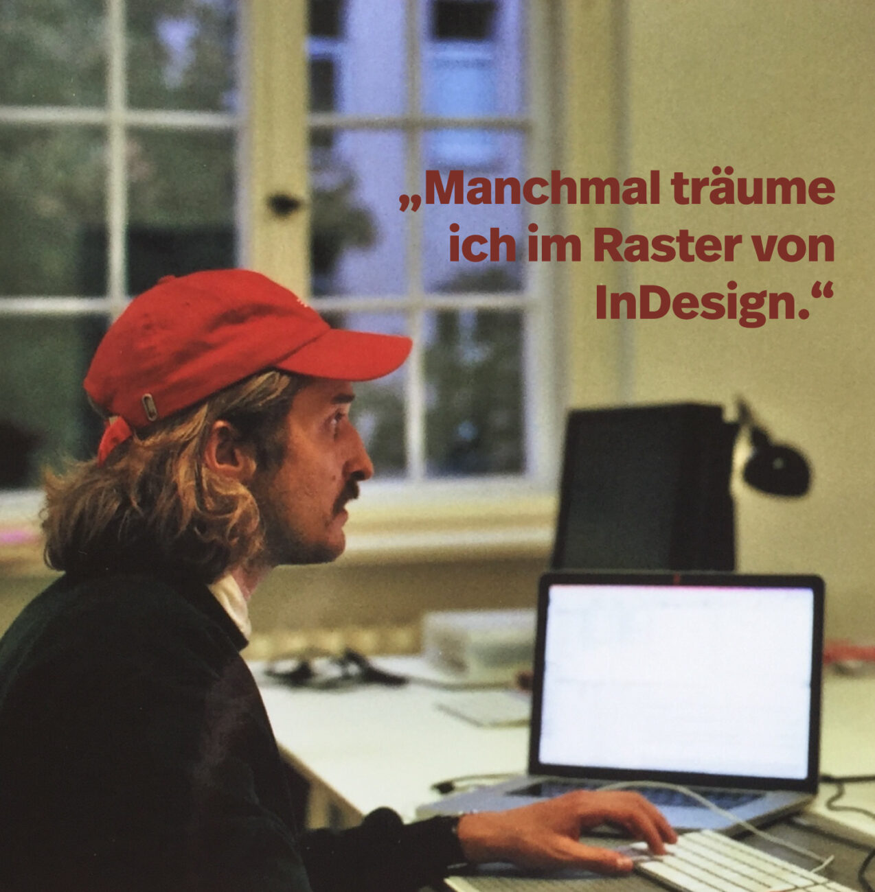Eine Person mit langen Haaren und einer roten Mütze sitzt an einem Schreibtisch und arbeitet an einem Laptop. Der Hintergrund zeigt ein Fenster und Büroartikel. Der deutsche Text auf dem Bild lautet: "Manchmal träume ich im Raster von InDesign.".