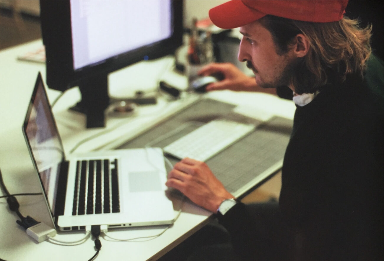 Eine Person mit einer roten Mütze arbeitet an einem Schreibtisch mit einem Laptop und einem großen Monitor, wobei sie eine Maus und eine Tastatur benutzt. Auf dem Schreibtisch sind Büromaterial und Kabel zu sehen.
