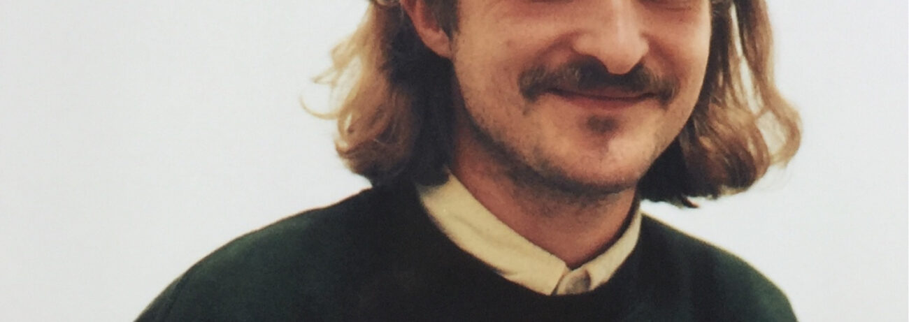 Eine Person mit schulterlangem hellbraunem Haar, Schnurrbart und einem leichten Lächeln, die einen dunklen Pullover über einem hellen Hemd mit Kragen trägt, steht vor einem schlichten hellen Hintergrund.