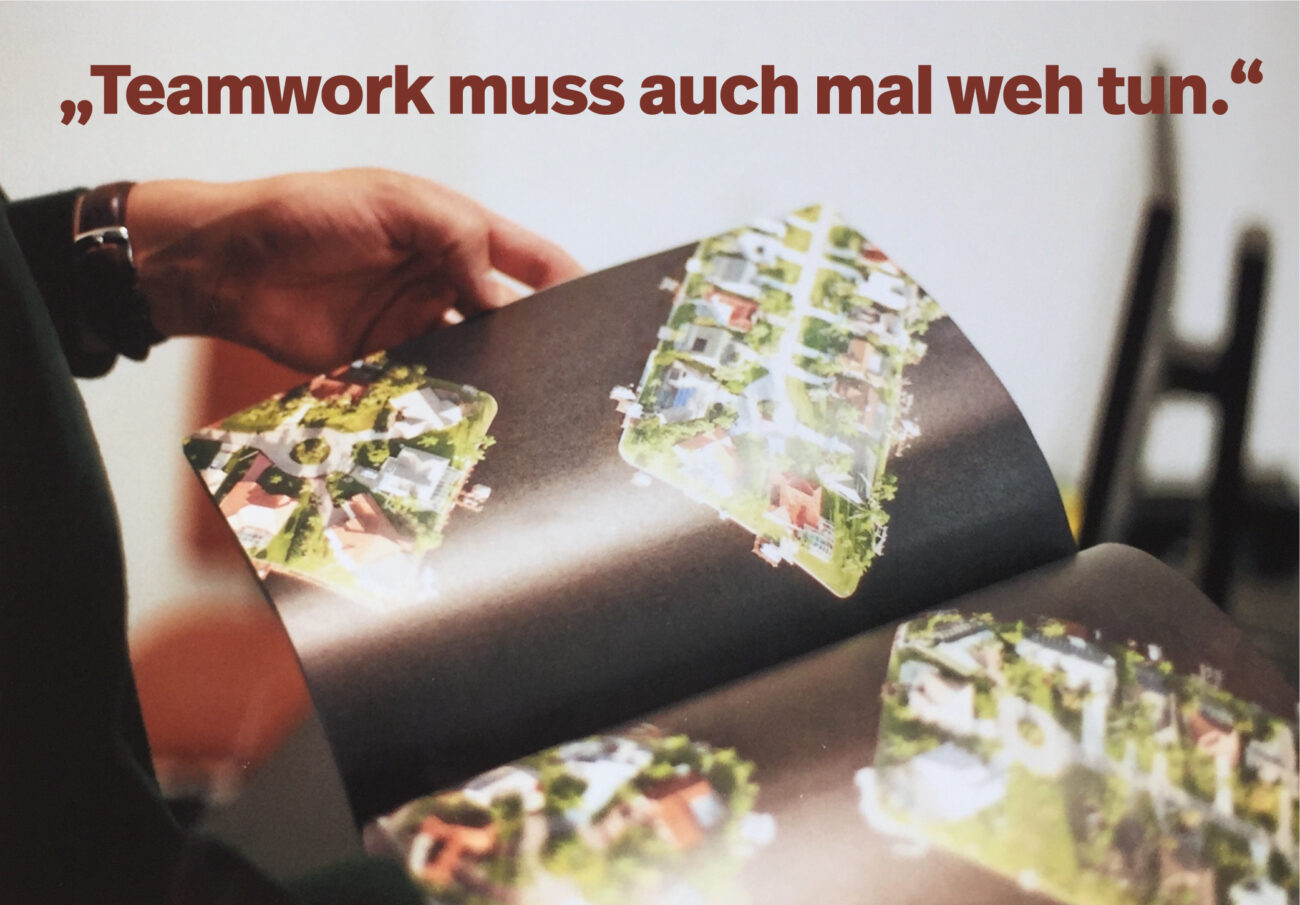 Eine Person hält ein aufgeschlagenes Magazin in der Hand, das Luftaufnahmen eines Wohngebiets zeigt. Oben steht ein deutscher Text: Teamwork muss auch mal weh tun. (Teamarbeit muss auch mal weh tun.).
