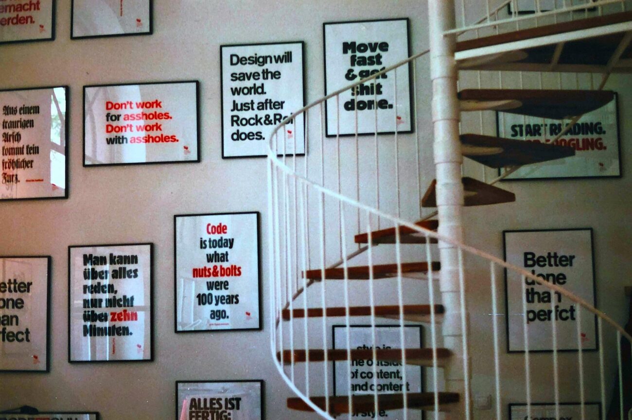 Eine weiße Wendeltreppe vor einer Wand, die mit gerahmten Motivationspostern mit fetten schwarzen und roten Texten in Englisch und Deutsch bedeckt ist.