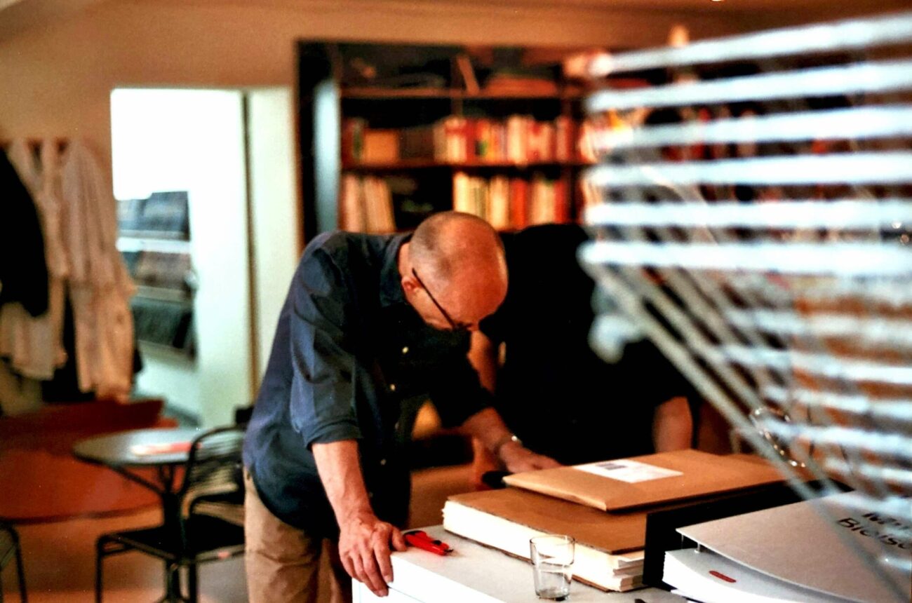Ein Mann mit Brille beugt sich über einen mit Papieren und Ordnern bedeckten Schreibtisch in einem gemütlichen, mit Büchern gefüllten Raum. Im Vordergrund sind Jalousien und ein Glas Wasser zu sehen.