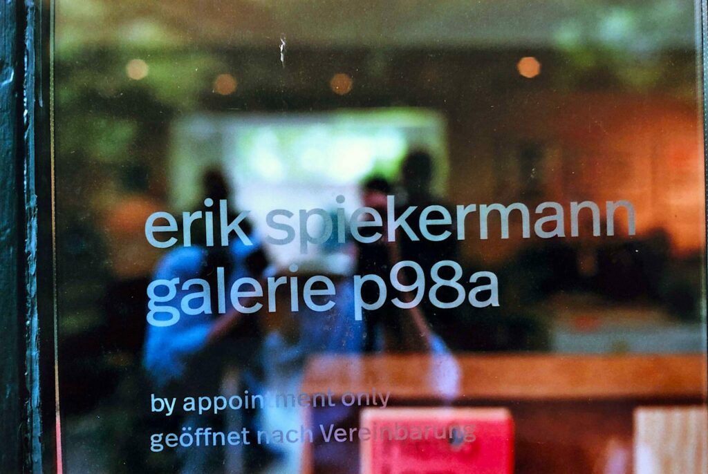 Nahaufnahme einer Glastür mit dem Text: erik spiekermann galerie p98a in Kleinbuchstaben und darunter nur nach Vereinbarung geöffnet, mit einer verschwommenen Innenraumszene, die sich im Glas spiegelt.