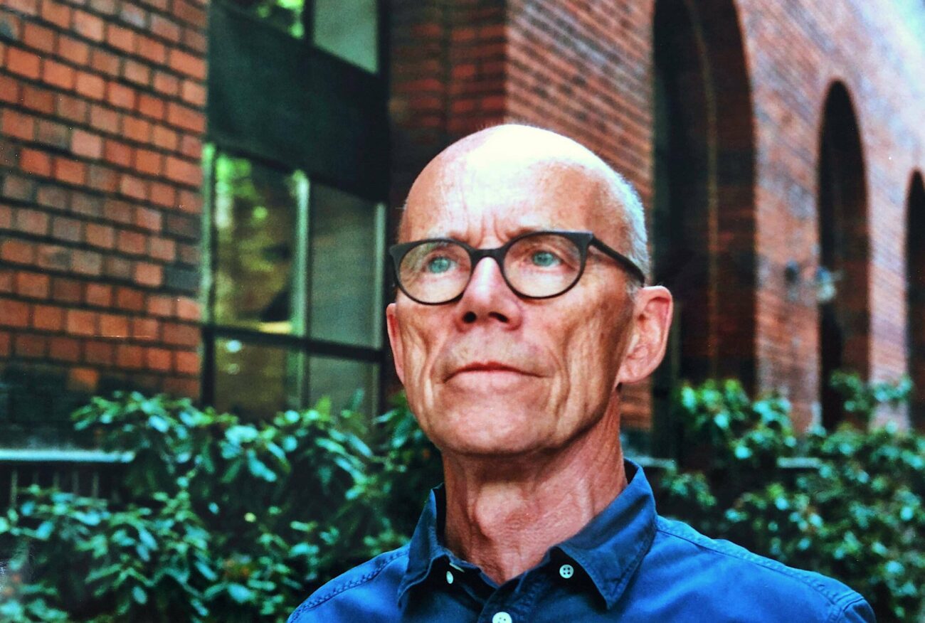 Ein Mann mittleren Alters mit Brille und blauem Hemd steht im Freien vor einem Backsteingebäude und grünen Büschen und blickt mit nachdenklicher Miene leicht nach oben.