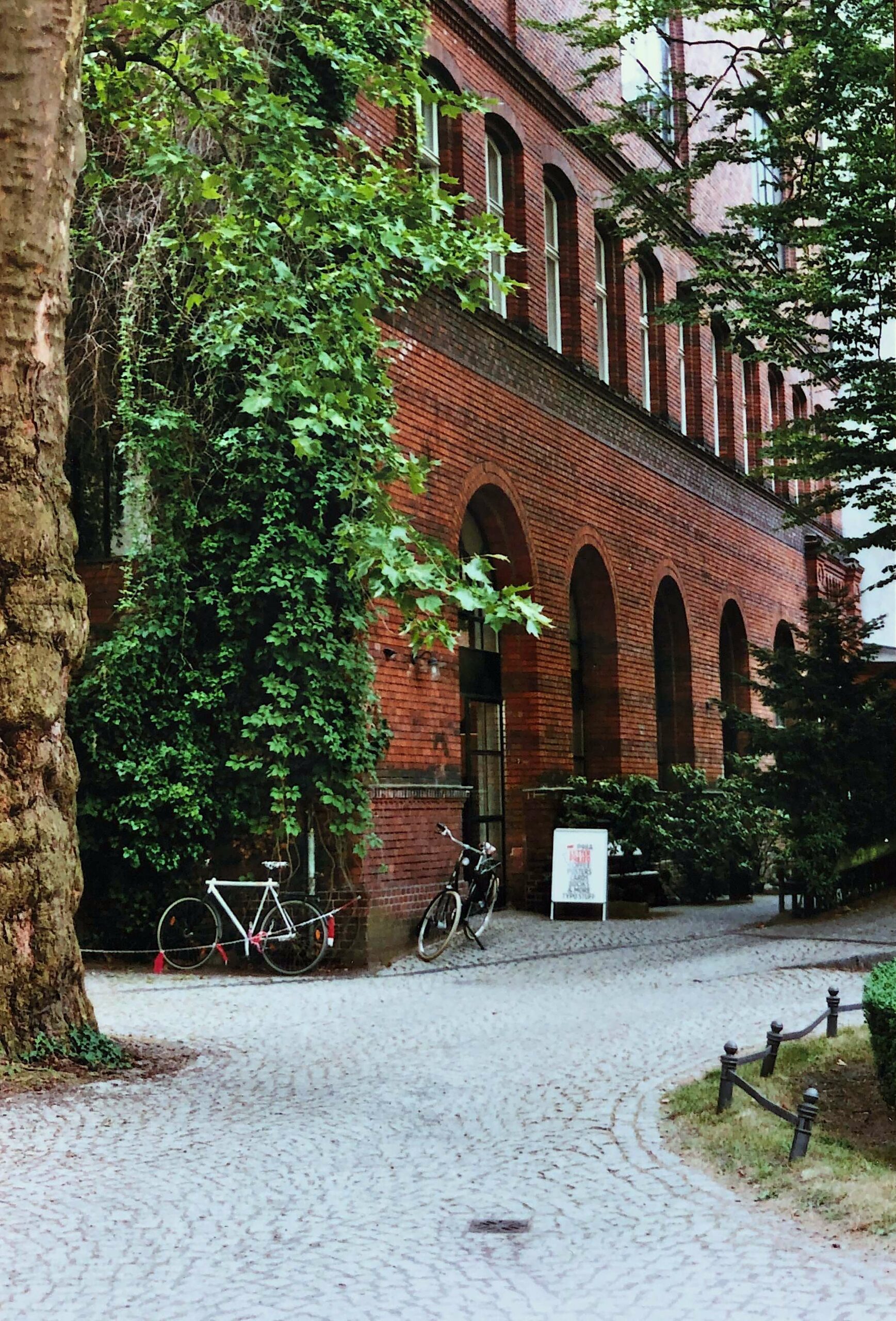 Ein kopfsteingepflasterter Weg schlängelt sich an einem roten Backsteingebäude mit großen Bogenfenstern vorbei. Zwei Fahrräder sind in der Nähe von efeubewachsenen Mauern geparkt, und belaubte Bäume umgeben die friedliche, schattige Szene.