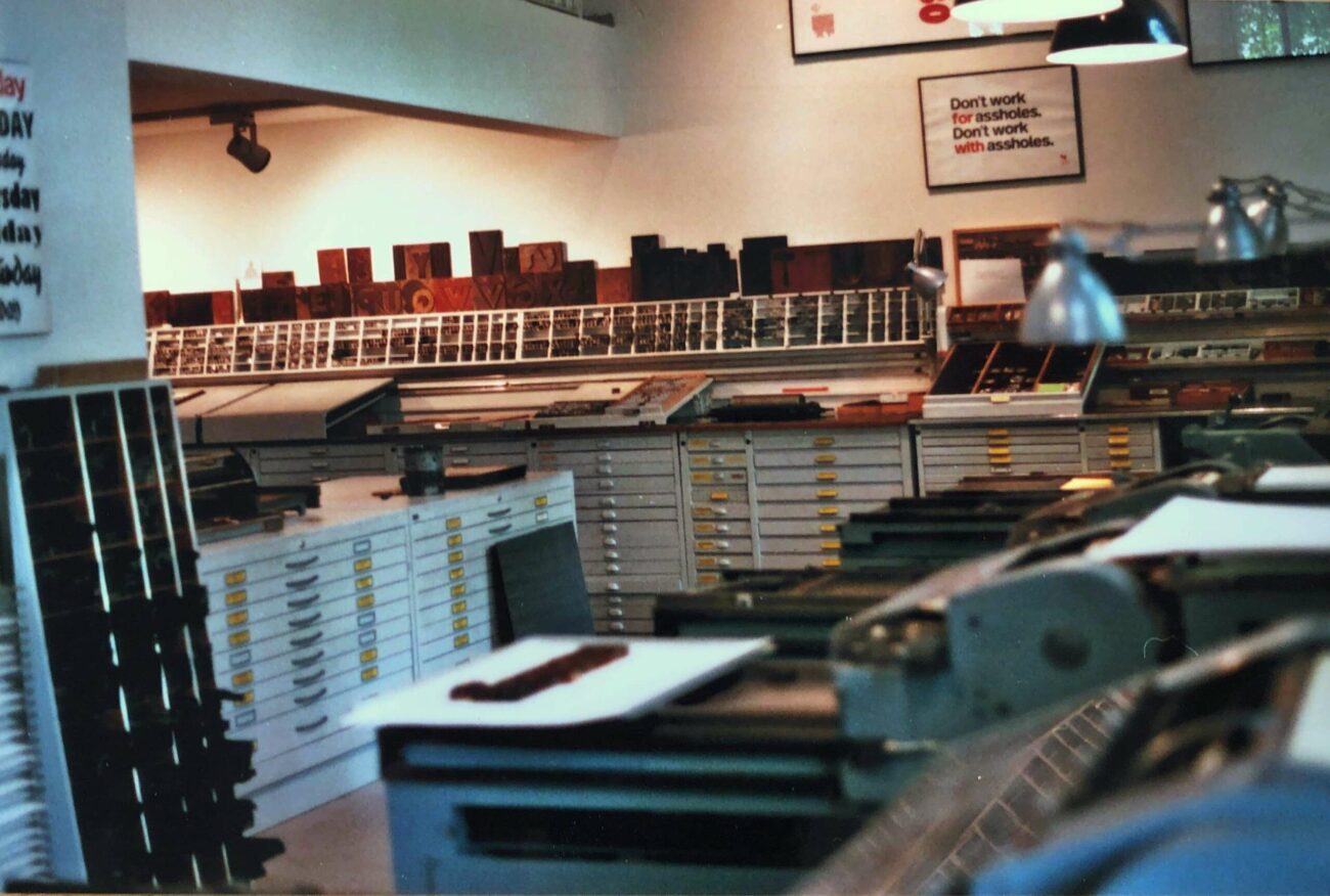 Eine Druckerei mit geordneten Schubladen und Regalen, gefüllt mit Druckstöcken und Schrift. Oberlichter erhellen den Arbeitsbereich. An der Wand hängt ein Schild mit der Aufschrift: "Arbeite nicht für Arschlöcher. Arbeite nicht mit Arschlöchern.".