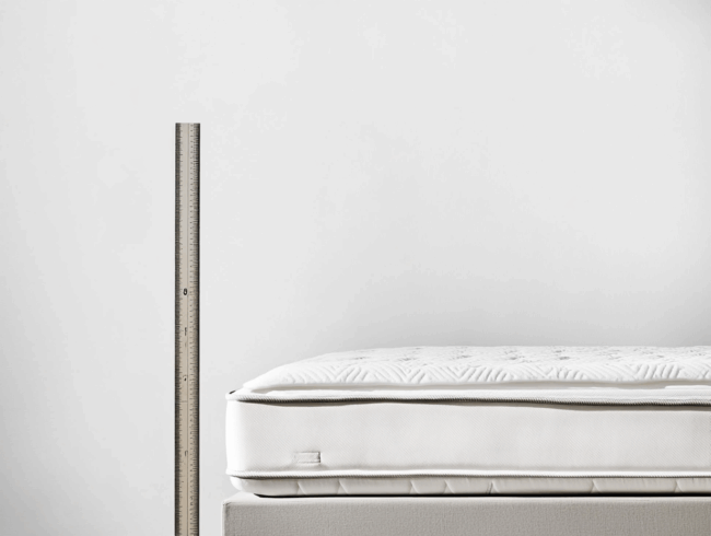 Ein Metalllineal steht aufrecht neben dem Rand einer weißen Matratze mit strukturierter Oberfläche und zeigt deutlich die Matratzenhöhe vor einem schlichten weißen Hintergrund.