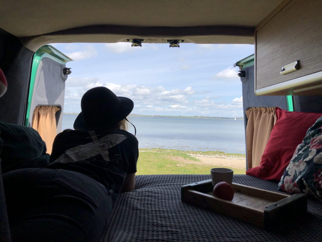Eine Person mit Hut liegt auf dem Rücksitz eines Lieferwagens und blickt auf einen ruhigen See oder ein Meer unter einem wolkenverhangenen Himmel, in der Nähe stehen ein Tablett, eine Tasse und ein Apfel. Die Türen des Wagens sind geöffnet und geben den Blick auf Gras und Wasser frei.