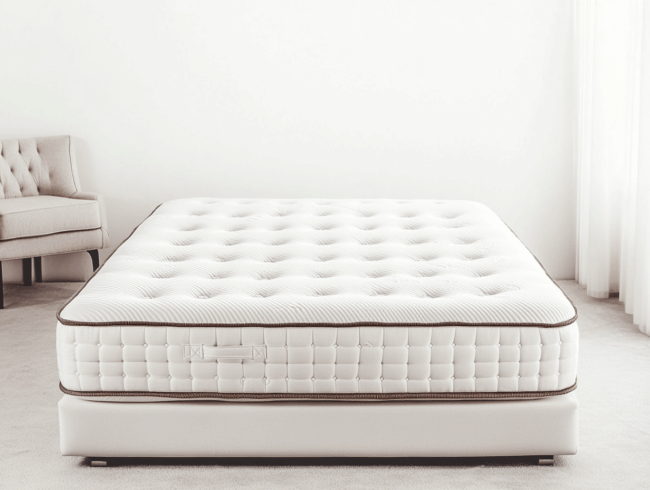 Ein ordentlich gemachtes Bett mit einer dicken, weißen, gesteppten Matratze - ideal, wenn man Matratze wenden möchte - steht auf einem weißen Lattenrost in einem hellen, minimalistischen Schlafzimmer. Im Hintergrund steht ein helles Sofa, und ein Fenster auf der rechten Seite ist mit transparenten Vorhängen verhängt.