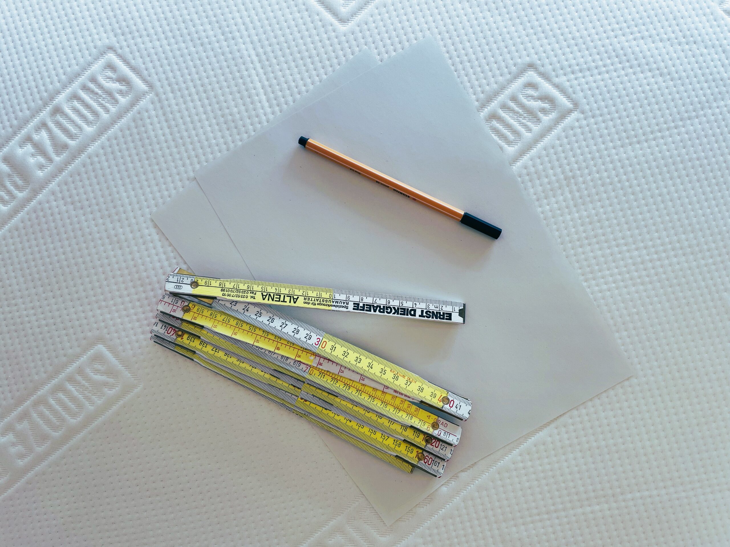 Zwei leere Blätter Papier, ein orangefarbener Stift mit schwarzer Kappe und ein gefalteter Messstab werden auf eine weiße, strukturierte Oberfläche mit einem sich wiederholenden SNÖZZE-Muster gelegt.