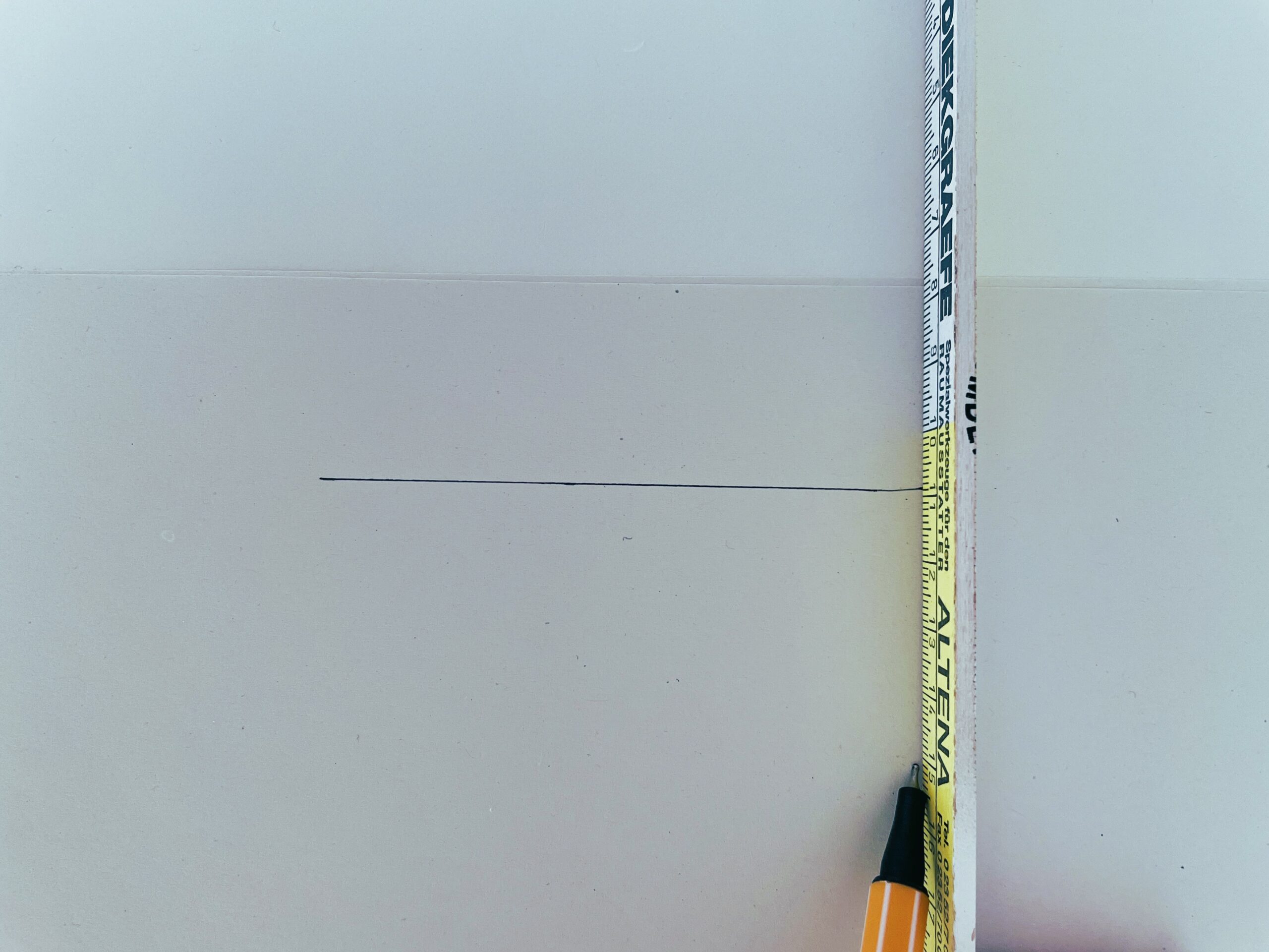 Ein Maßband, das senkrecht neben einer horizontalen, mit Bleistift gezogenen Linie auf einer weißen Fläche gehalten wird, wobei das Maßband die Höhe der Linie markiert.