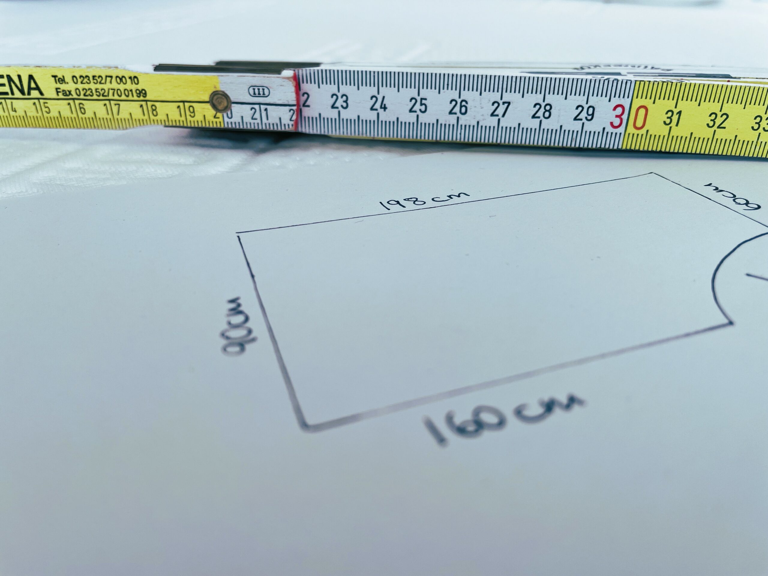 Nahaufnahme eines Maßbandes, das über eine handgezeichnete rechteckige Skizze auf Papier gespannt ist, die mit den Maßen 198 cm, 160 cm und 90 cm beschriftet ist. Das Maßband zeigt Zahlen in Zentimetern an.