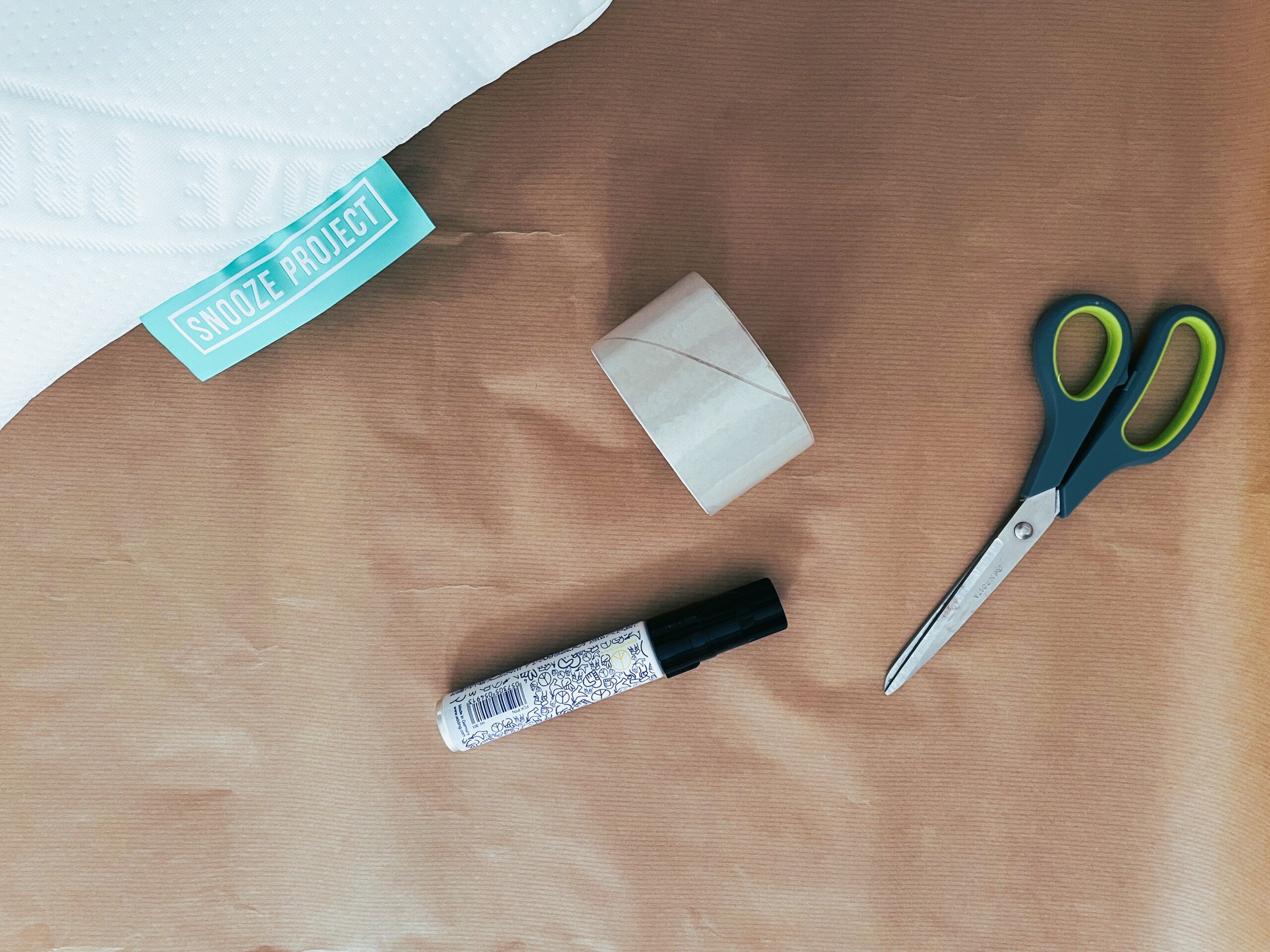 Eine Schere, ein Permanentmarker und eine Rolle Klebeband liegen auf braunem Packpapier neben der Ecke eines weißen Kissens mit der Aufschrift Snooze Project.