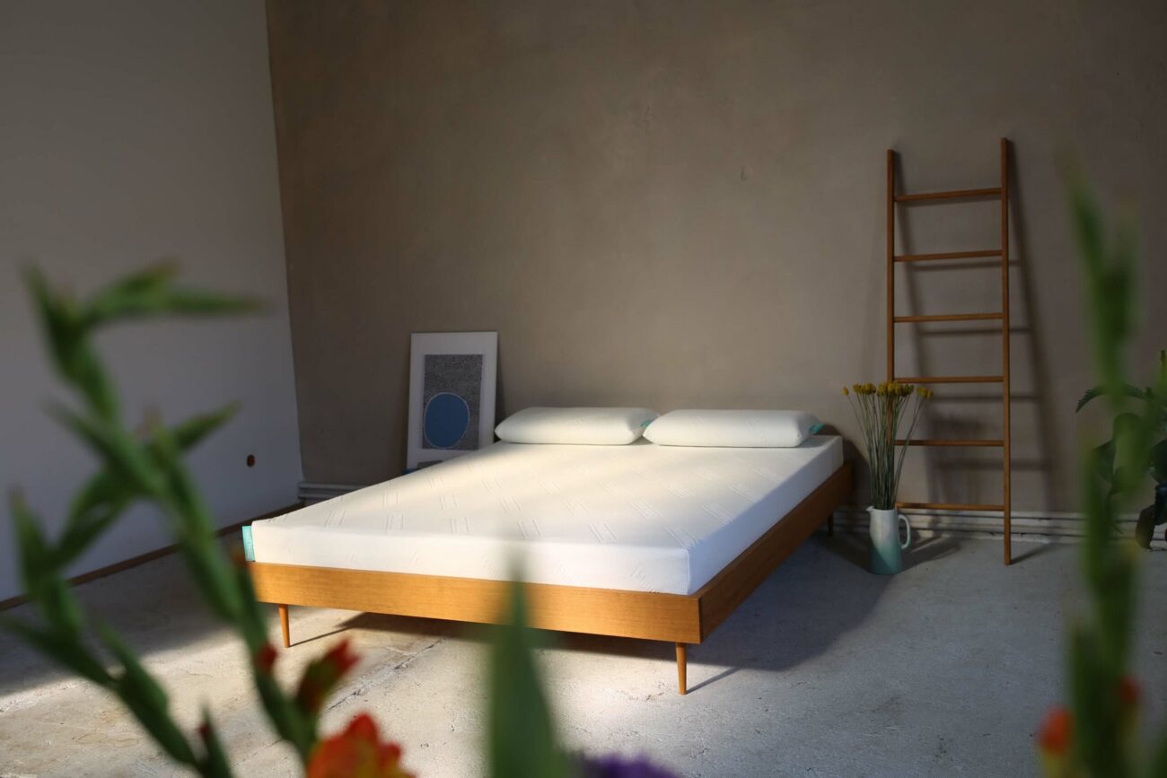 A minimalist bedroom with a wooden bed frame, two pillows and a white mattress. In the corner there is a wooden ladder, a framed work of art, a vase of yellow flowers and blurred plants in the foreground.