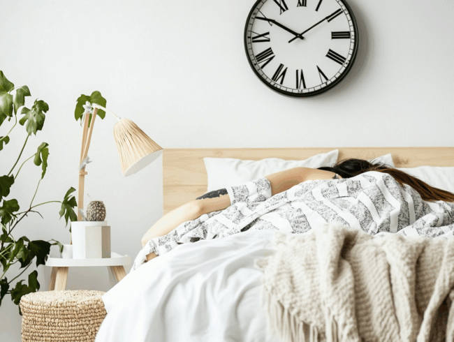 Eine Person schläft in einem Bett mit weißer und gemusterter Bettwäsche und genießt einen erholsamen Schlafzyklus. Eine große Uhr an der Wand zeigt 10:10. Auf dem Nachttisch stehen eine Lampe, Bücher und eine kleine Pflanze, und in der Ecke steht eine Blattpflanze.