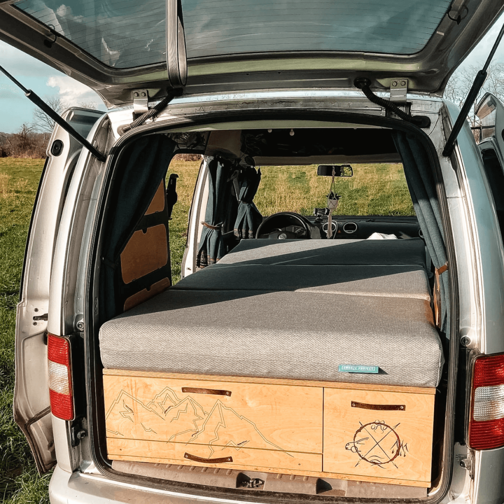 A camper van with an open back door showing a neatly made bed with gray bedding and wooden drawers underneath, parked in a meadow with a blue sky in the background.