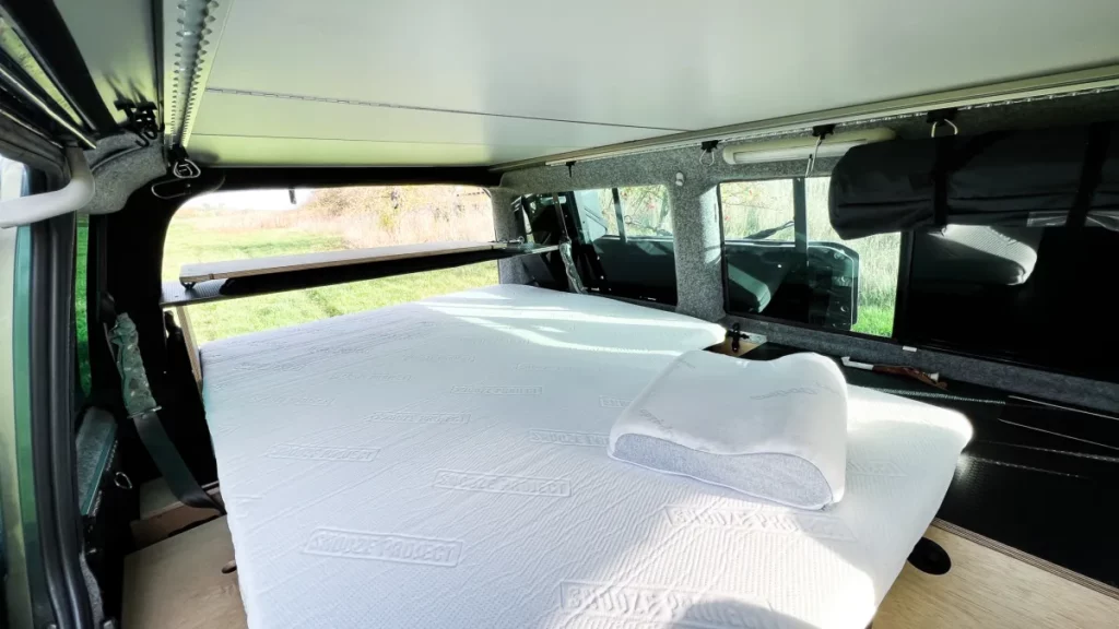A neatly made bed with a pillow inside a camper van with large windows, sunlight streaming in and green grass outside. The interior seems compact and efficiently furnished for sleeping.
