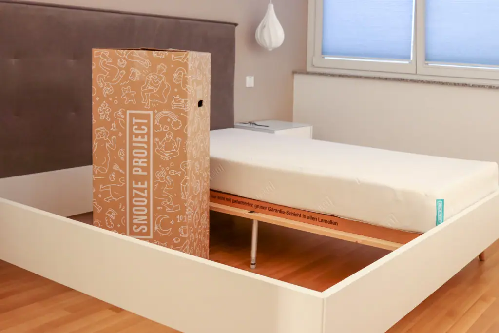 A wrapped mattress labeled Snooze Project stands next to an unwrapped mattress on a white bed frame in a modern bedroom with a wooden floor and a large window.