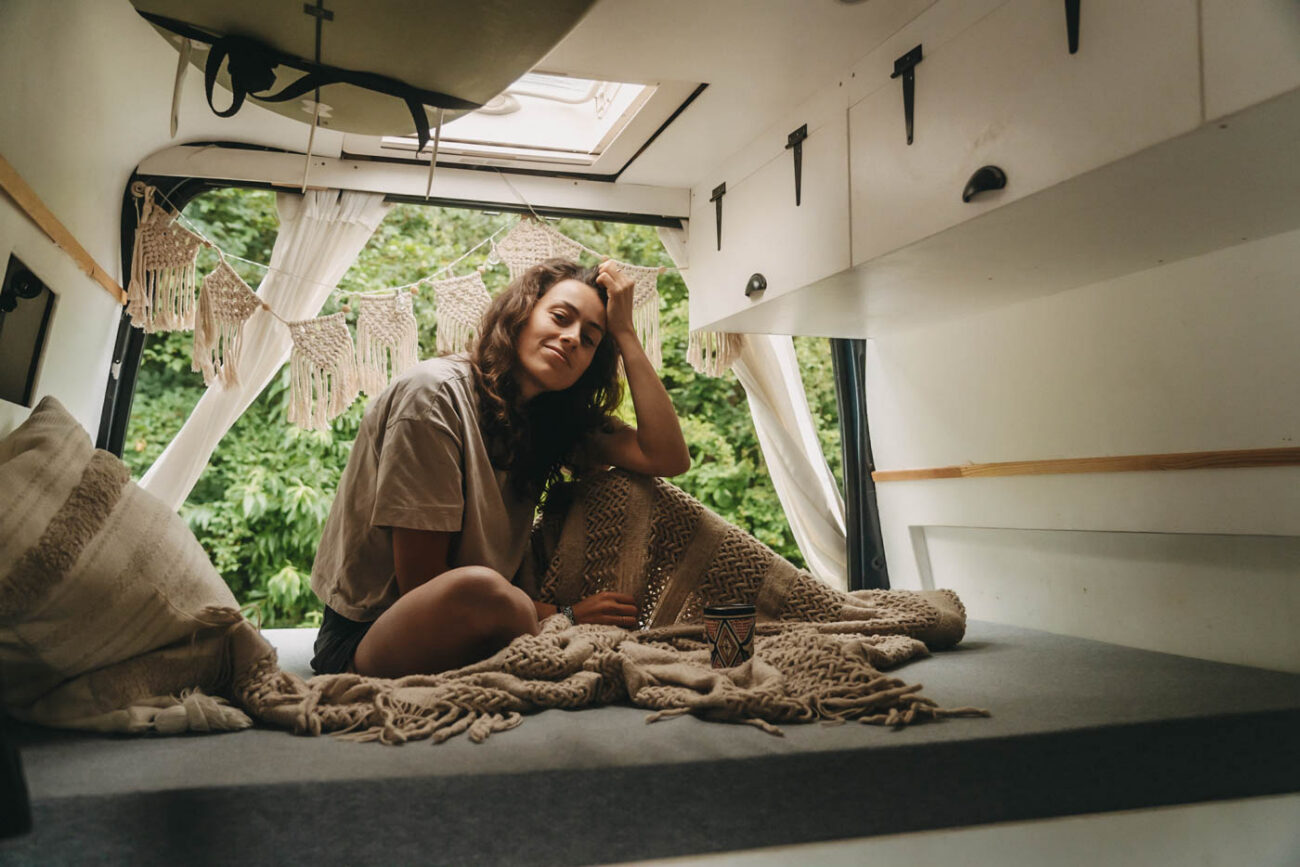 A woman sits on a bed in a comfortably furnished mobile home, surrounded by blankets and cushions, while sunlight streams in through a skylight and green plants can be seen in front of the open door.