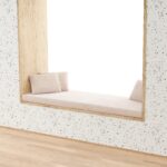 Ein gemütlicher Fensterplatz mit einem Rechteckigen Sitzfenster Polster nach Maß und zwei passenden Kissen ist in eine terrazzomäßig gemusterte Wand eingebaut und überblickt einen hellen Raum mit Holzfußboden.