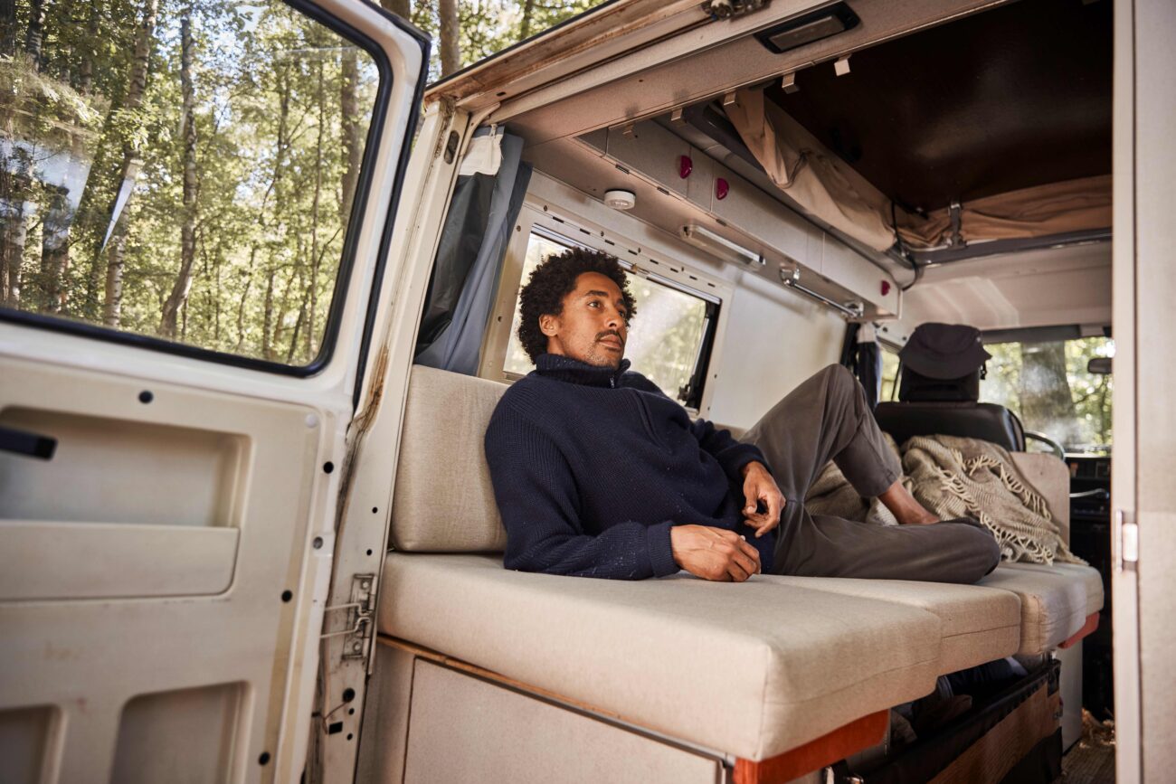 Ein Mann liegt auf einer gepolsterten Snooze Project-Bank in einem Wohnmobil mit offener Seitentür und blickt nach draußen. Das Sonnenlicht fällt durch die hohen Bäume im Hintergrund. Der Innenraum wirkt gemütlich und entspannt - ein perfekter, von DHDL inspirierter Rückzugsort.