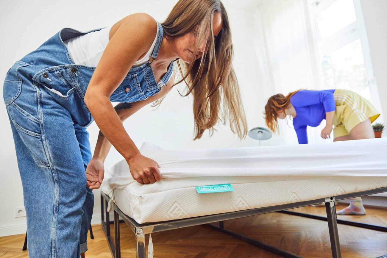 Zwei Frauen machen zusammen ein Bett in einem hellen Raum. Eine zieht das Laken auf der Matratze zurecht, während die andere das Bettzeug glättet. Das Sonnenlicht fällt durch ein nahe gelegenes Fenster ein.