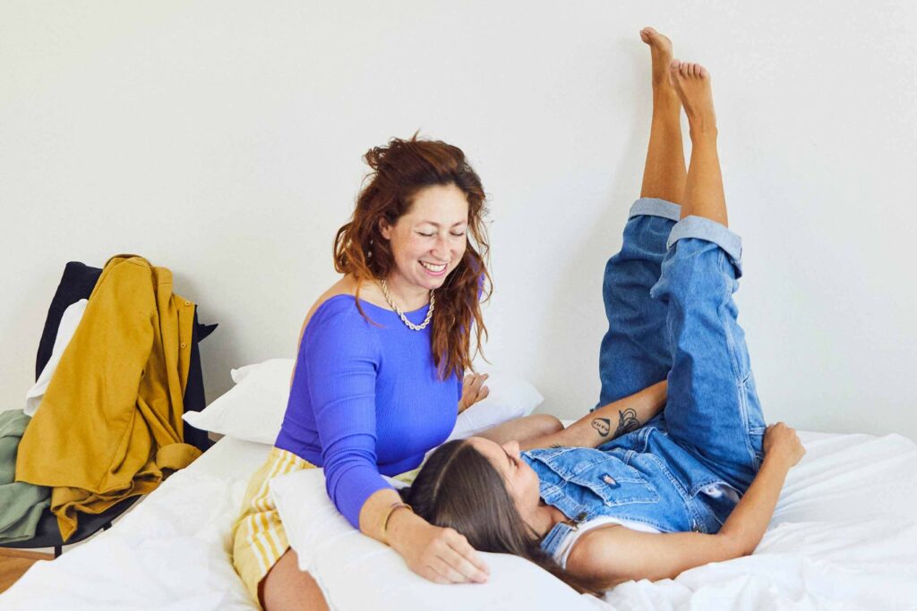 Zwei Frauen entspannen sich auf einem Bett, lächeln und lachen zusammen. Die eine sitzt in einem blauen Top, die andere liegt mit den Beinen an der Wand und trägt einen Jeans-Overall. Ein gelber Mantel hängt an einem Stuhl im Hintergrund.