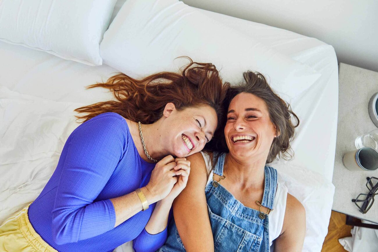 Zwei Frauen liegen auf einem Bett, lachen und lächeln sich an. Die eine trägt ein blaues langärmeliges Hemd, die andere einen Jeans-Overall. Ihre Hände sind ineinander verschränkt, und sie wirken fröhlich und entspannt.