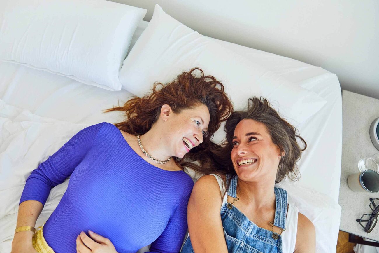 Zwei Frauen liegen nebeneinander auf einem Bett, lächeln sich an und schauen sich an. Die eine trägt ein blaues Oberteil, die andere einen Jeans-Overall. Die Szene wirkt entspannt und fröhlich.