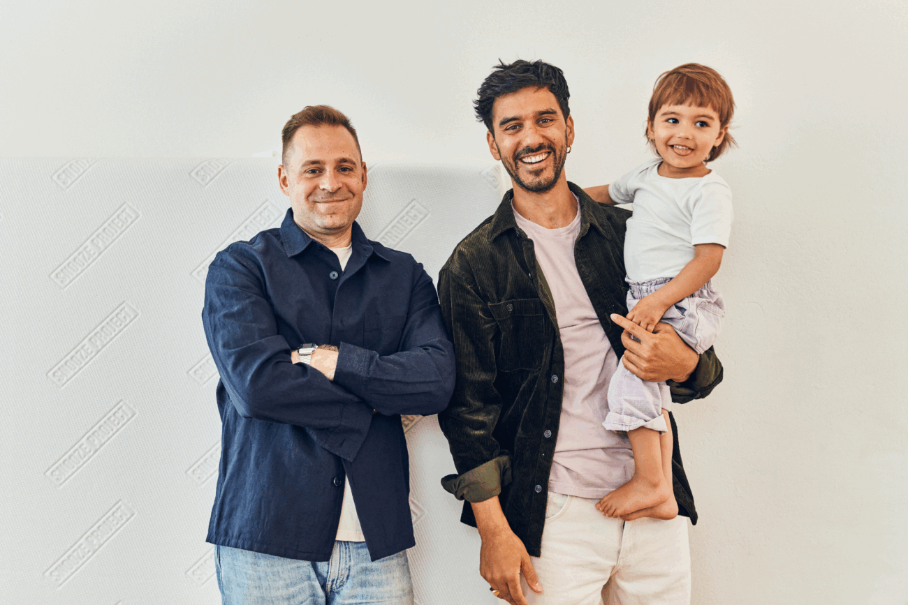 Zwei lächelnde Männer stehen nebeneinander vor einer hellen Wand; einer hält ein kleines Kind, das lächelt, während der andere mit verschränkten Armen dasteht. Alle wirken entspannt und glücklich.