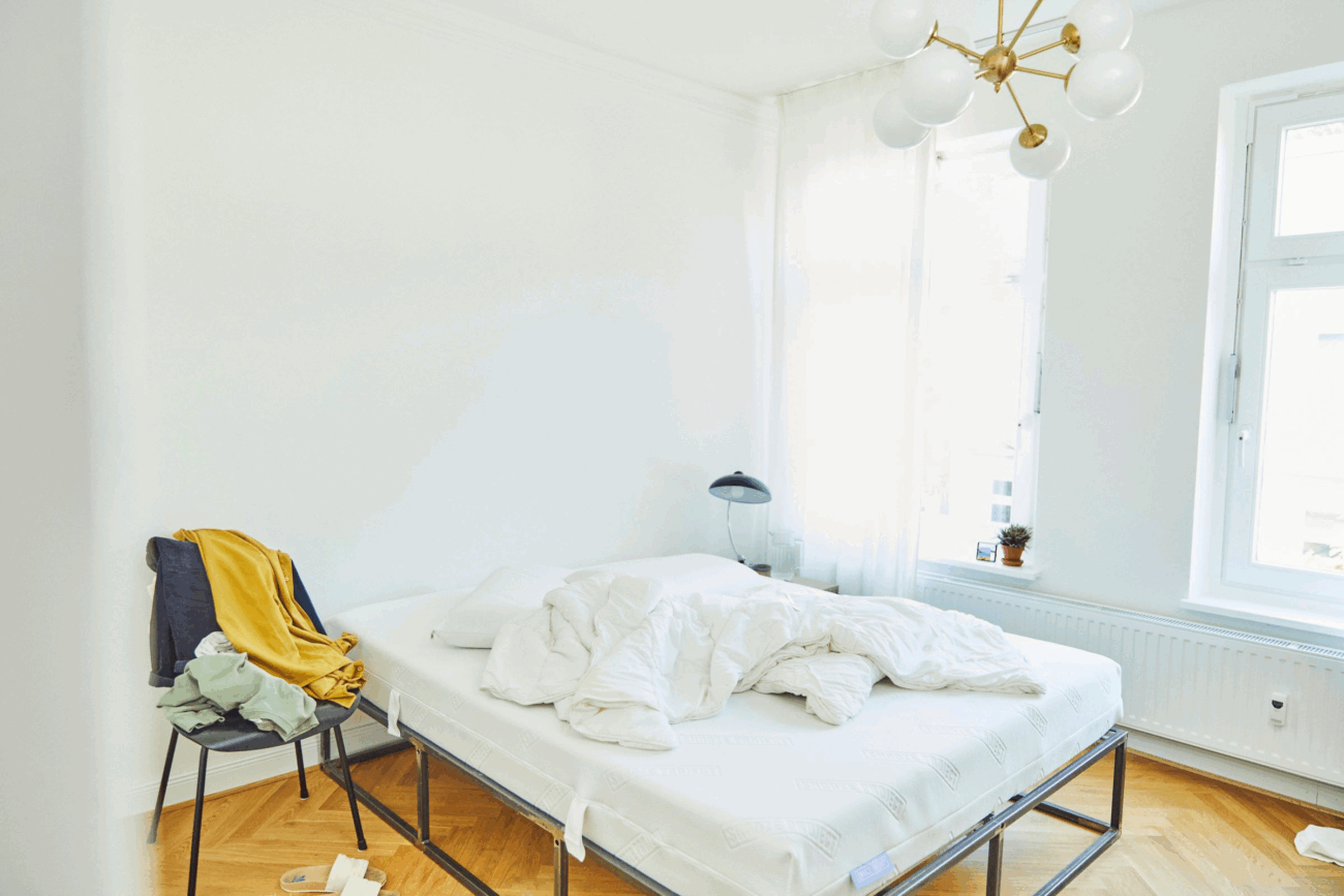 Ein helles, minimalistisches Schlafzimmer mit weißen Wänden und großen Fenstern. Ein ungemachtes Bett mit weißem Bettzeug steht neben einem Stuhl mit Kleidung. Ein moderner Kronleuchter hängt von der Decke und eine kleine Pflanze steht auf der Fensterbank.