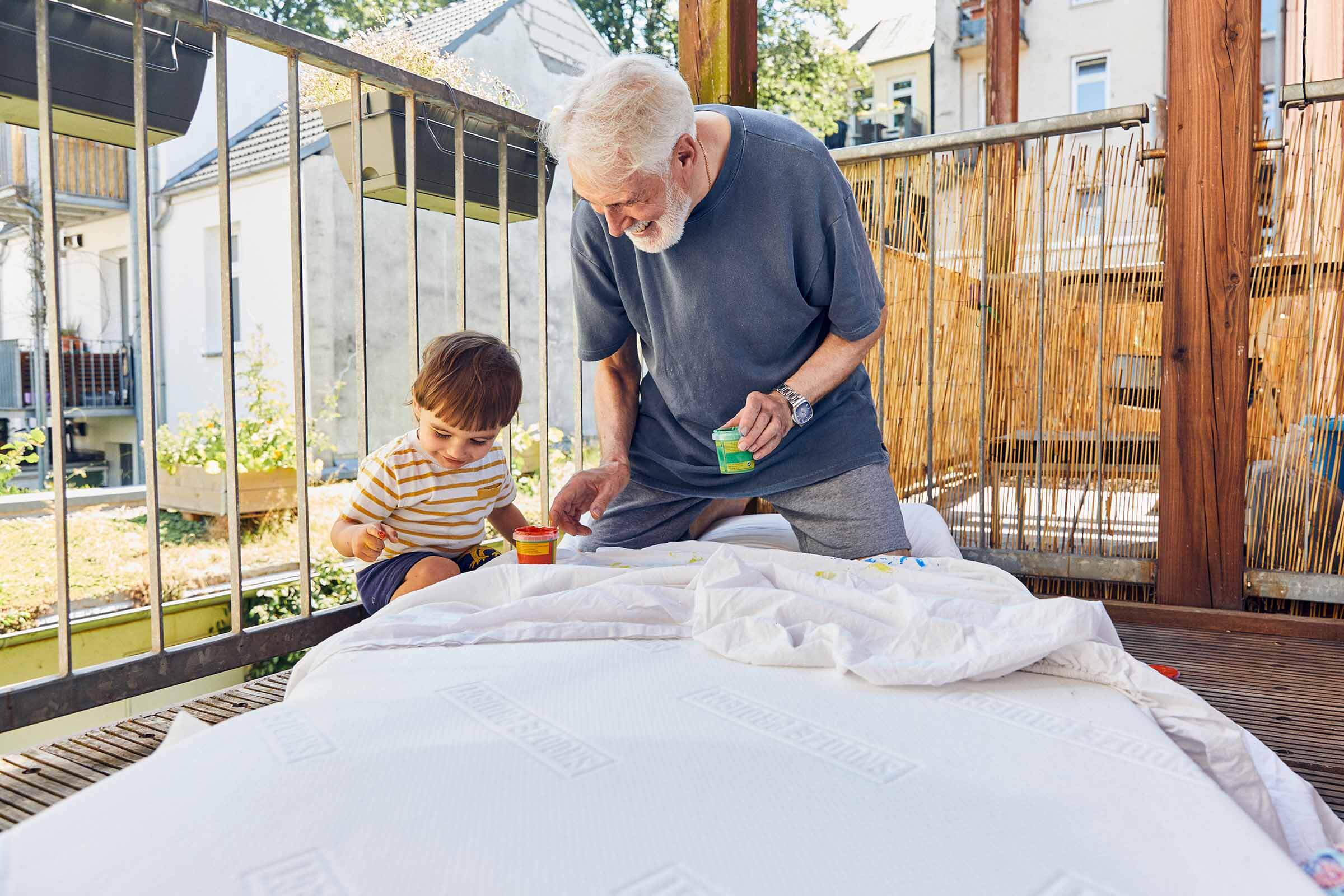 Ein älterer Mann und ein kleines Kind sitzen im Freien auf einer Holzterrasse und malen gemeinsam. Sie konzentrieren sich auf ihre Tätigkeit, umgeben von Sonnenlicht und Grün, und legen ihre Malutensilien auf eine weiße Fläche vor sich.