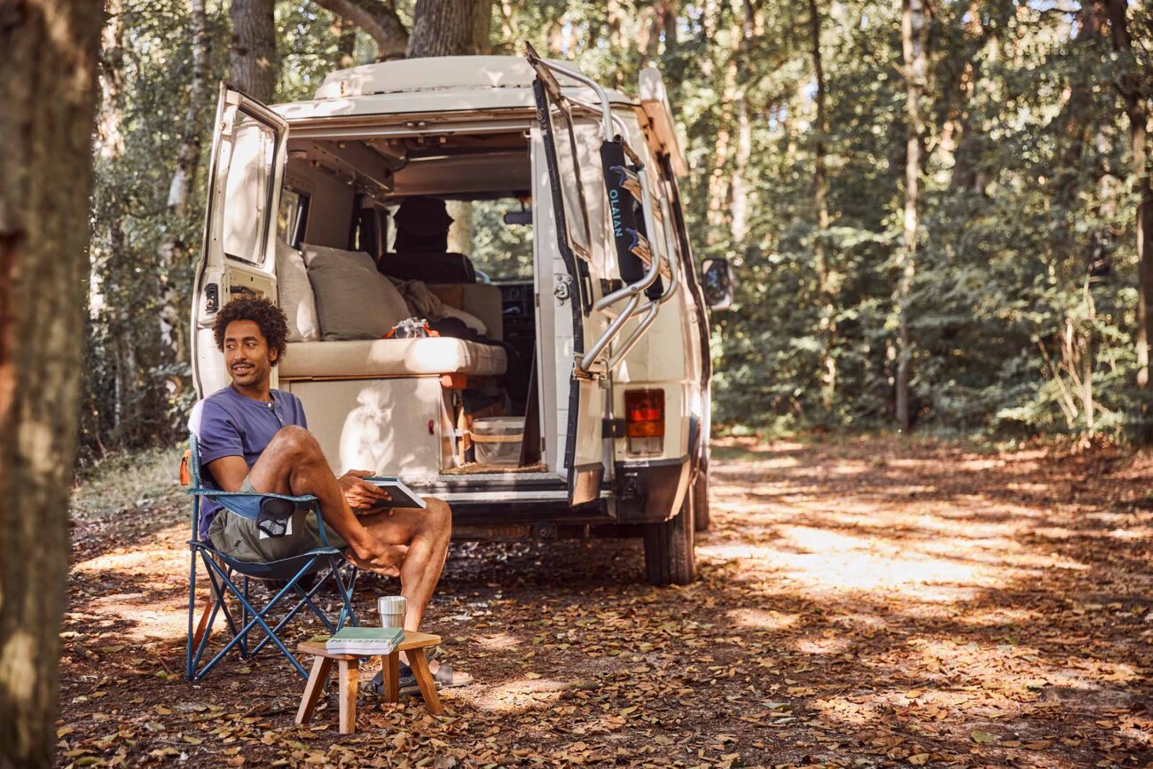 Eine Person sitzt in einem Campingstuhl neben einem offenen Wohnmobil in einem Wald und liest ein Buch. In der Nähe steht ein kleiner Tisch mit einem Buch und einer Tasse. Das Sonnenlicht fällt durch die Bäume auf den mit Blättern bedeckten Boden.
