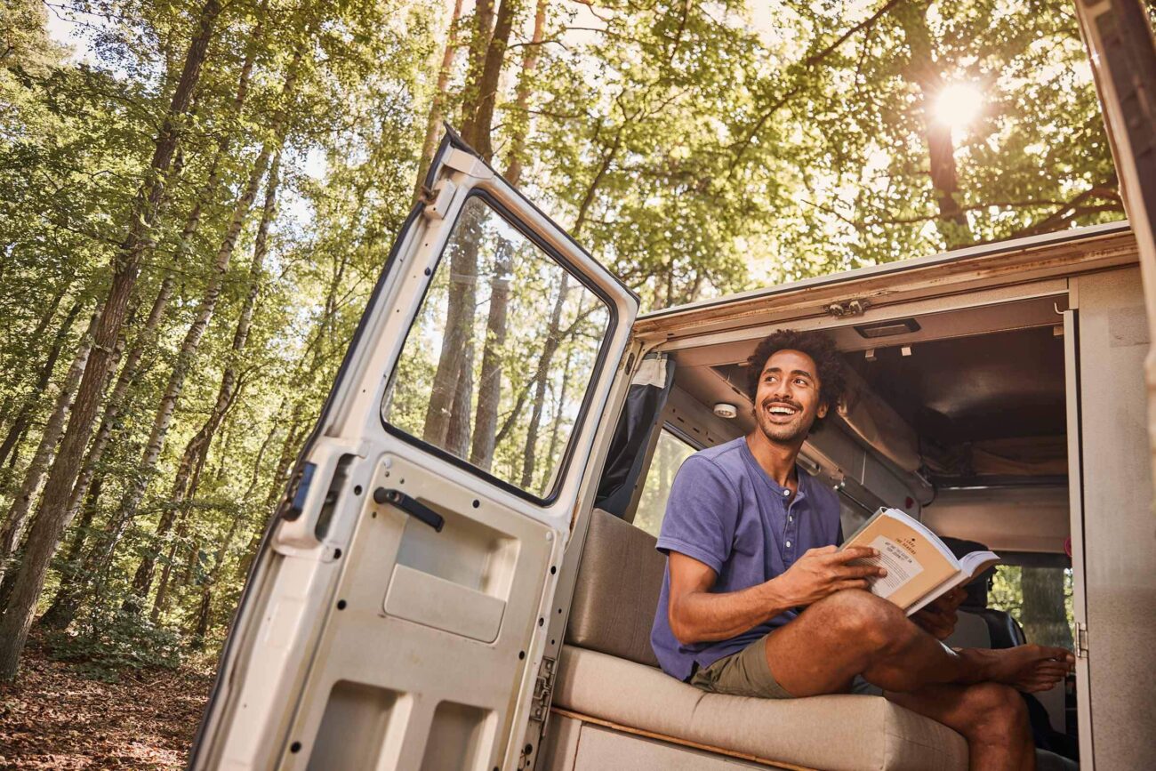 Ein Mann sitzt in der offenen Tür eines Wohnmobils, das in einem sonnenbeschienenen Wald geparkt ist, und lächelt, während er ein Buch liest. Das Sonnenlicht fällt durch die Bäume und schafft eine friedliche, entspannte Atmosphäre im Freien.
