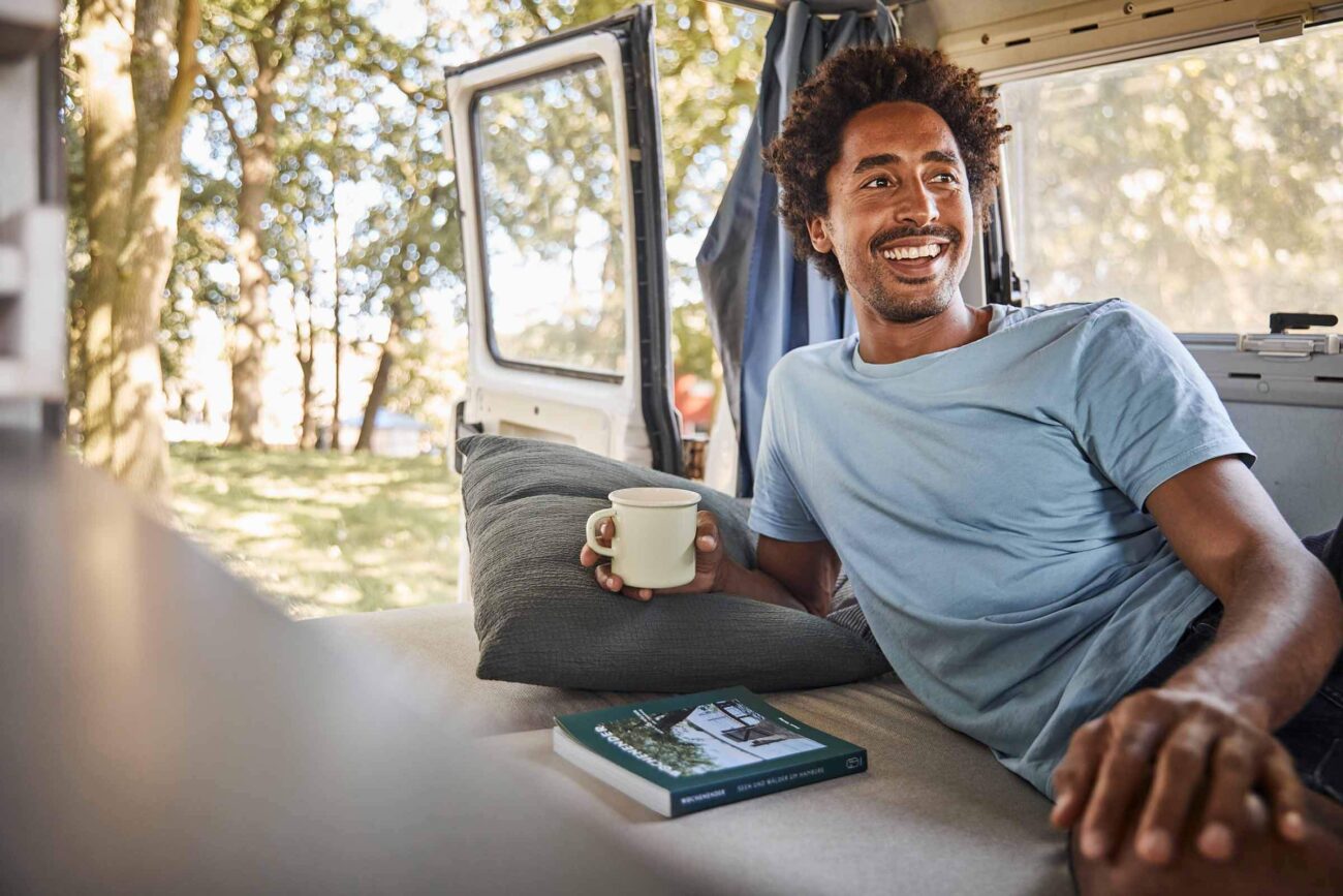 Ein Mann sitzt in einem Lieferwagen mit offener Tür, hält einen Becher in der Hand und lächelt. Er trägt ein hellblaues T-Shirt und ruht sich auf einem Kissen aus, neben dem er ein Buch liegen hat. Draußen sind Bäume und Sonnenlicht zu sehen.