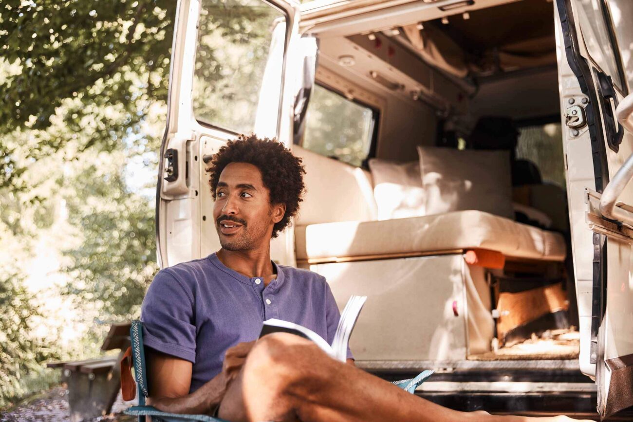 Eine Person sitzt auf einem Stuhl neben einem offenen Lieferwagen, hält ein Buch in der Hand und blickt zur Seite. Das Innere des Wagens ist zu sehen, mit einem Bett und Kissen. Das Sonnenlicht fällt durch die Bäume im Hintergrund.