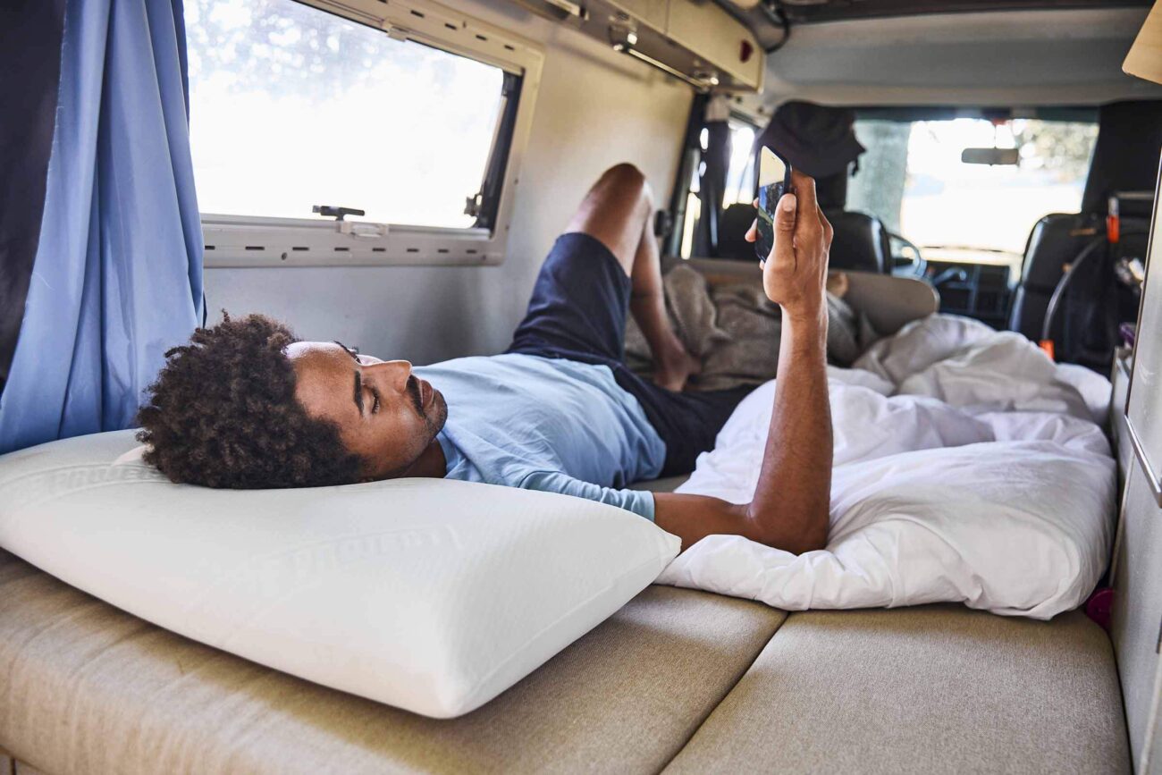 Eine Person liegt auf einem Bett in einem Wohnmobil, stützt sich auf ein weißes Kissen und eine Decke und schaut auf ein Smartphone. Durch ein Fenster strömt Sonnenlicht herein, und das Innere des Wohnmobils ist um sie herum zu sehen.