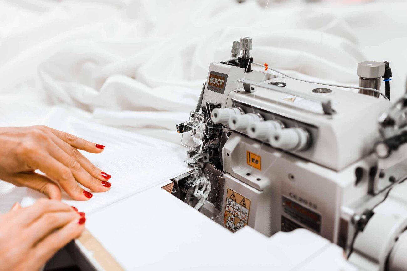 Nahaufnahme von Händen mit rotem Nagellack, die weißen Stoff durch eine industrielle Nähmaschine führen, mit weißem Stoff im Hintergrund.