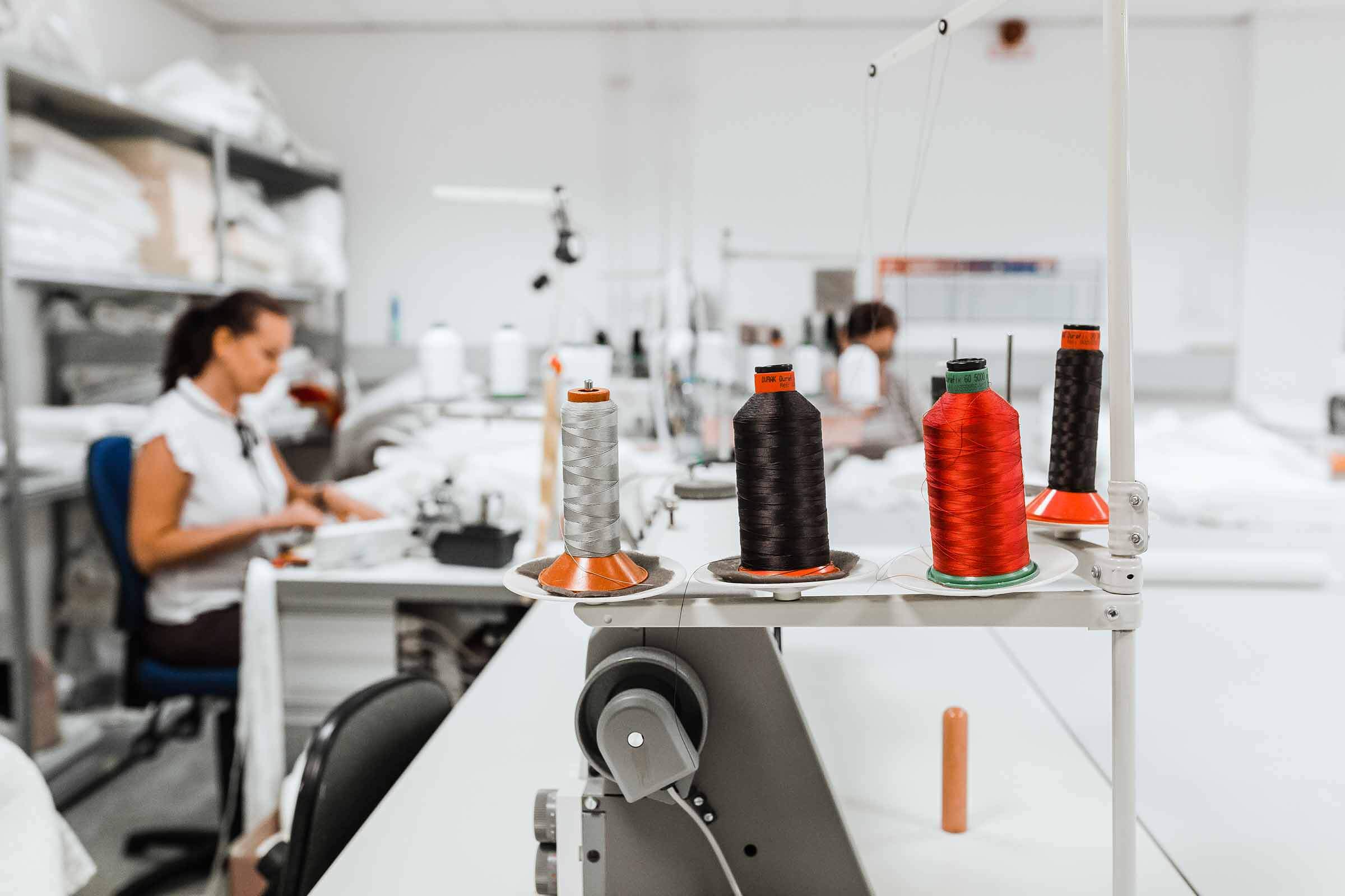 Fadenspulen in verschiedenen Farben auf einer Nähmaschine im Vordergrund, im Hintergrund zwei Personen, die an Schreibtischen in einer hellen, organisierten Textilwerkstatt arbeiten.