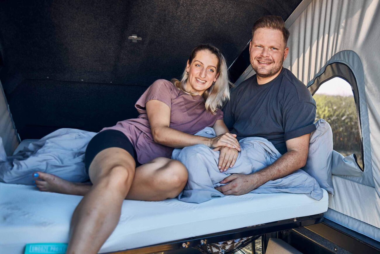 Ein lächelndes Paar sitzt zusammen auf einem Bett in einem Wohnmobil, lehnt sich an Kissen und hält sich an den Händen, wirkt entspannt und glücklich in einem gemütlichen, sonnendurchfluteten Raum.