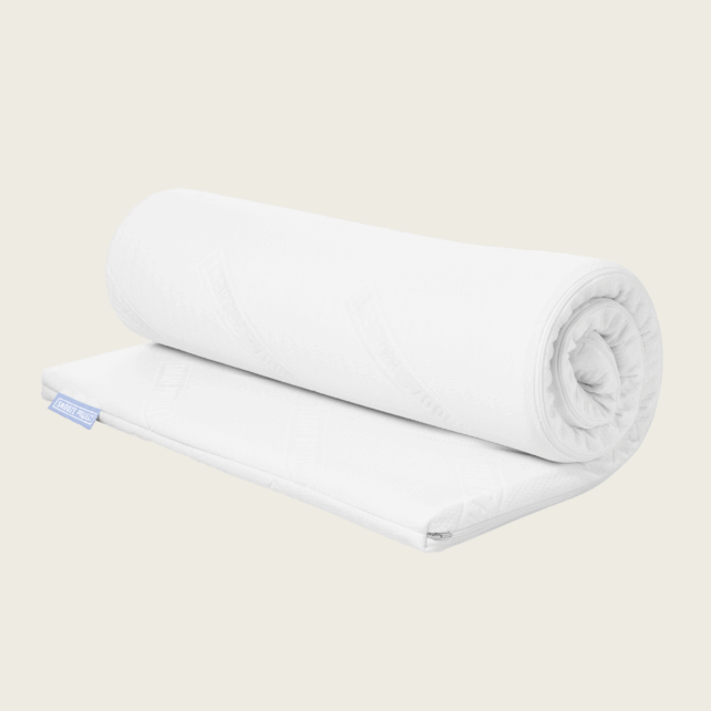 Der Familienbett Topper, eine weiße Matratzenauflage mit einem Stoffetikett an einer Ecke, ist ordentlich aufgerollt und steht vor einem schlichten, hellen Hintergrund.