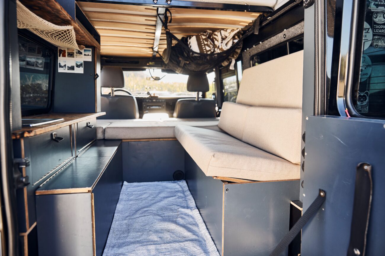 Innenraum eines Wohnmobils mit einer gepolsterten Sitzbank - perfekt zum einfachen Reinigen der Polster -, kompakten Schränken, einem kleinen Teppich und einer Holzlattendecke. Durch die offenen Hecktüren fällt natürliches Licht in den Innenraum, der Fahrerbereich ist vorne sichtbar.