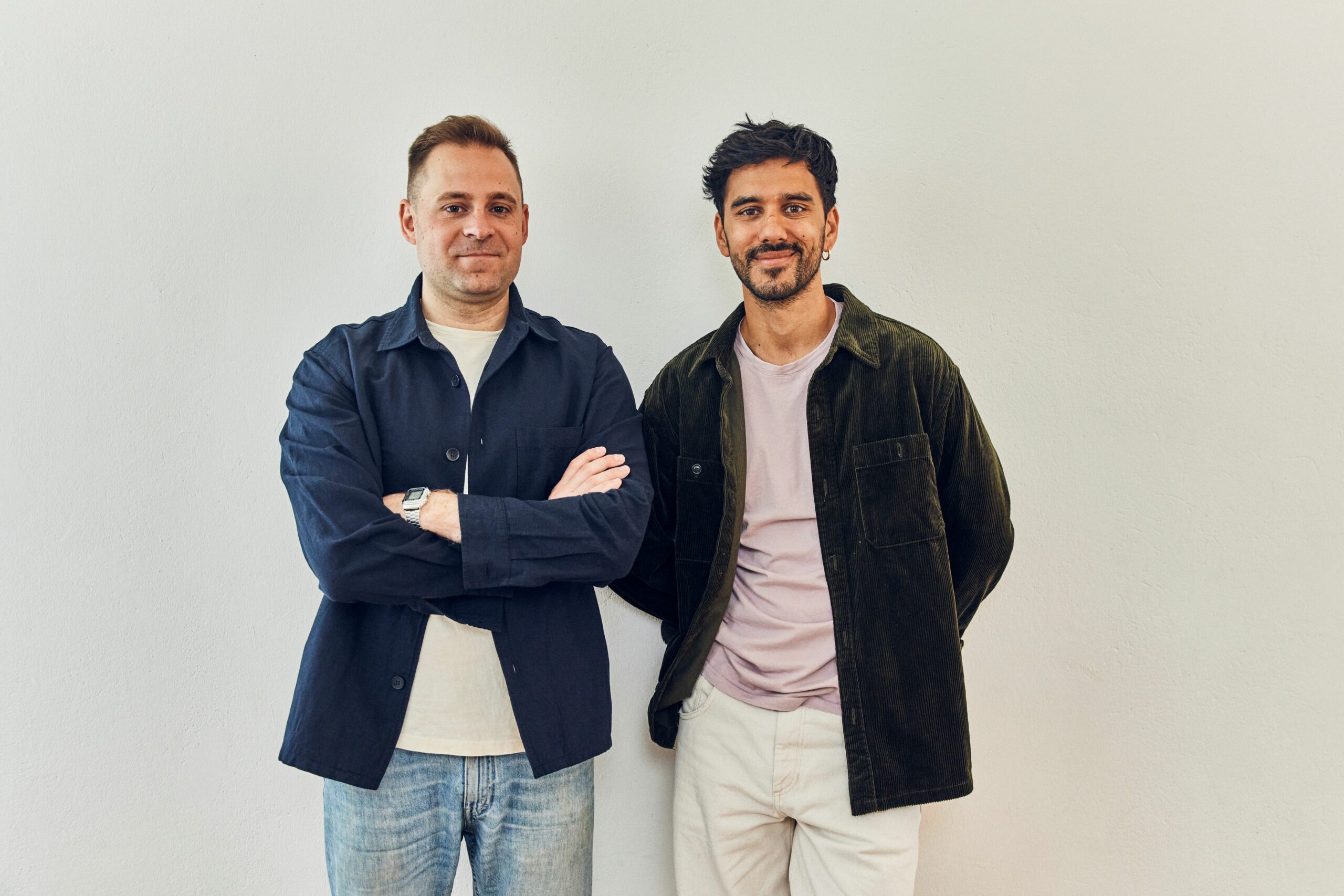 Zwei Männer stehen nebeneinander vor einer schlichten weißen Wand und lächeln. Der eine, in dunkler Jacke und Jeans, verschränkt die Arme; der andere, in dunkelgrüner Jacke und heller Hose, steht mit den Händen auf dem Rücken. Sie sind bereit, das Einrichtungskonzept des Snooze Project neu zu starten.
